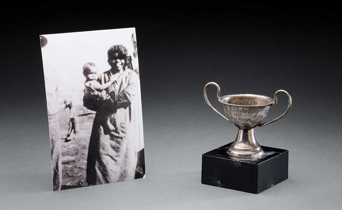 A sports trophy, in the form of a silver coloured metal cup with two handles, which is mounted on a square black support. An inscription on the cup reads 'N.K.S. Sub. Jnr Champ / 1972 / Andrea Mason'.