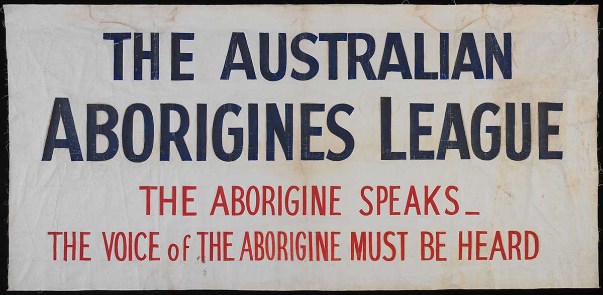 A rectangular calico banner featuring an inscription in blue and red paint which reads 'THE AUSTRALIAN ABORIGINES LEAGUE, THE ABORIGINE SPEAKS_THE VOICE of THE ABORIGINE MUST BE HEARD'. - click to view larger image