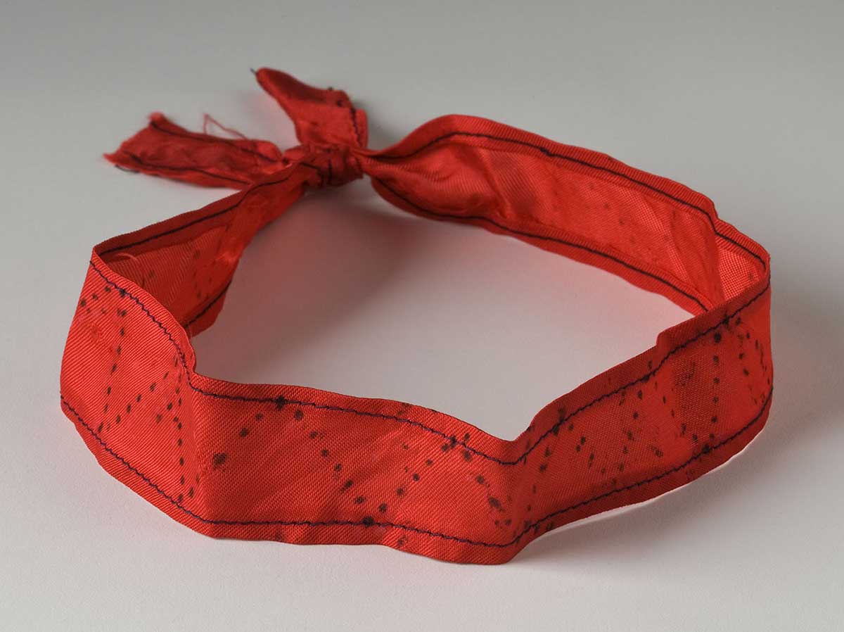 A headband fashioned from a length of silky red material, machine hemmed along both edges with thick black cotton thread and with the two ends joined together with a double knot. - click to view larger image