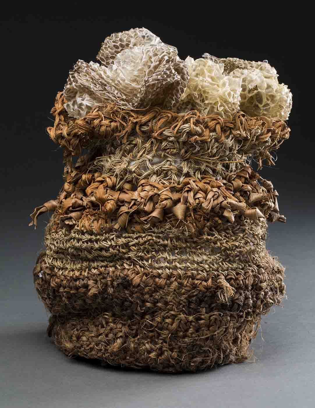 A vertical woven bag with a round base made of various types of fibres that are different shades of brown. Placed on top of the bag inside it is a long curled piece of snake skin which is also different shades of brown. - click to view larger image