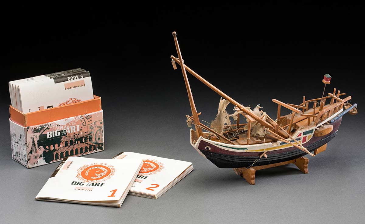 A model of a boat with two masts and cotton thread representing ropes. The boat is painted in red, white, green, yellow and black. The model is mounted onto a wooden base. Adjacent to the model is a box of miniature manuals.