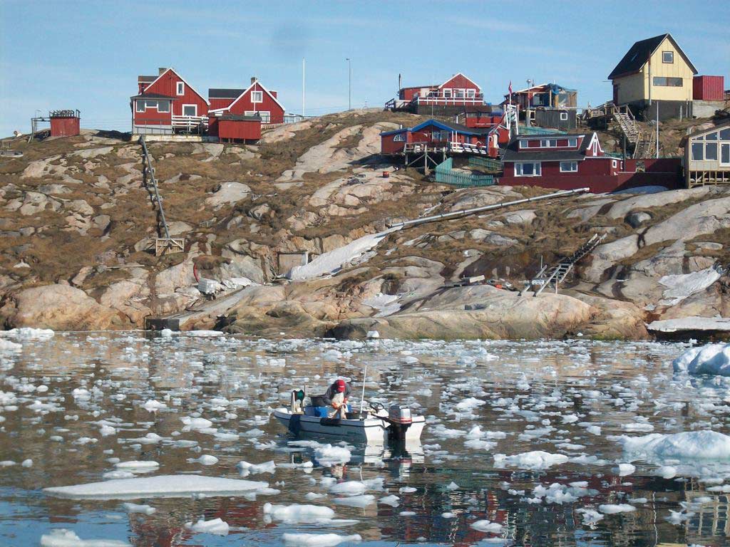 Coloured photograph of a man in a boat on an expanse of water close to a hilly shoreline. The water is covered with pieces of floating ice and there are houses on the hill in the background. - click to view larger image
