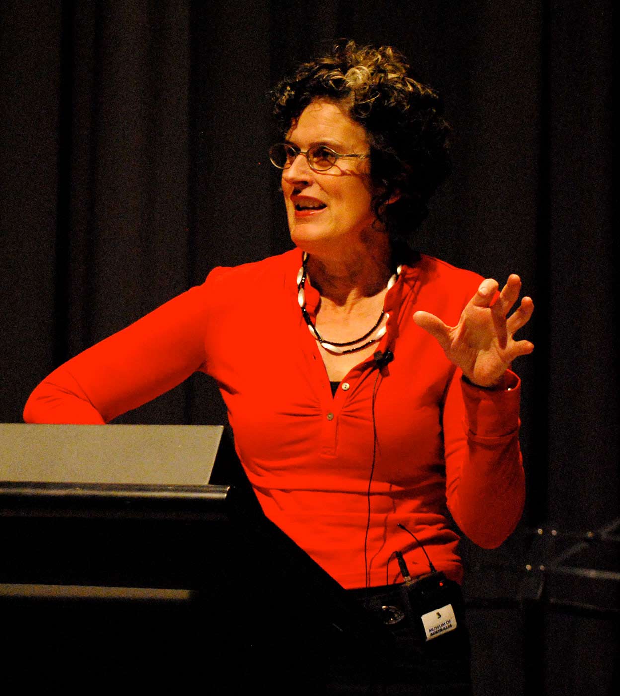 A woman wearing a red top stands at a lectern on stage in mid sentence. - click to view larger image