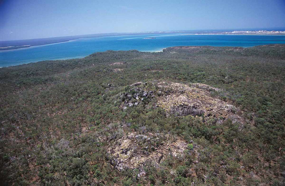 A colour photograph of a rocky outcrop on a hill. The outcrop is in the middle of the photograph, surrounded by partially bare earth. The hill is surrounded by a gently undulating plateau, which is covered in clumped vegetation. In the background is an area of blue open water. On the horizon is the edge of another landform.  - click to view larger image