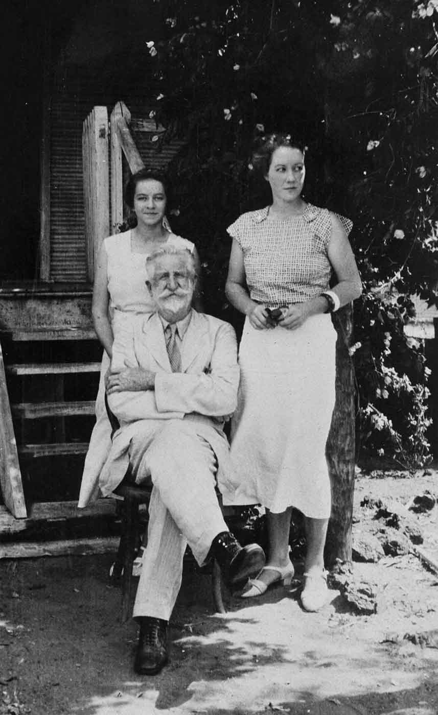 Black and white photo of two women standing beside a seated elderly man, at the entrance to an old wooden house.. - click to view larger image