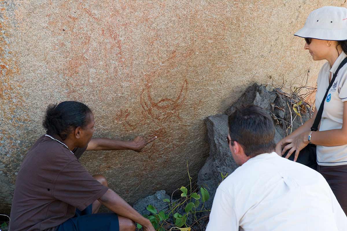 A woman  to paintings on a rock while two other people look on. - click to view larger image