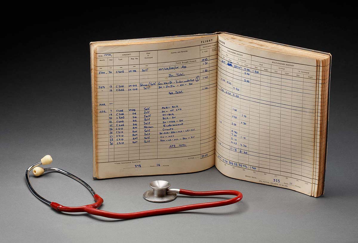A flight log book opened to show hand-written entries from 1974. A stethoscope with red plastic cover is beside the book. - click to view larger image