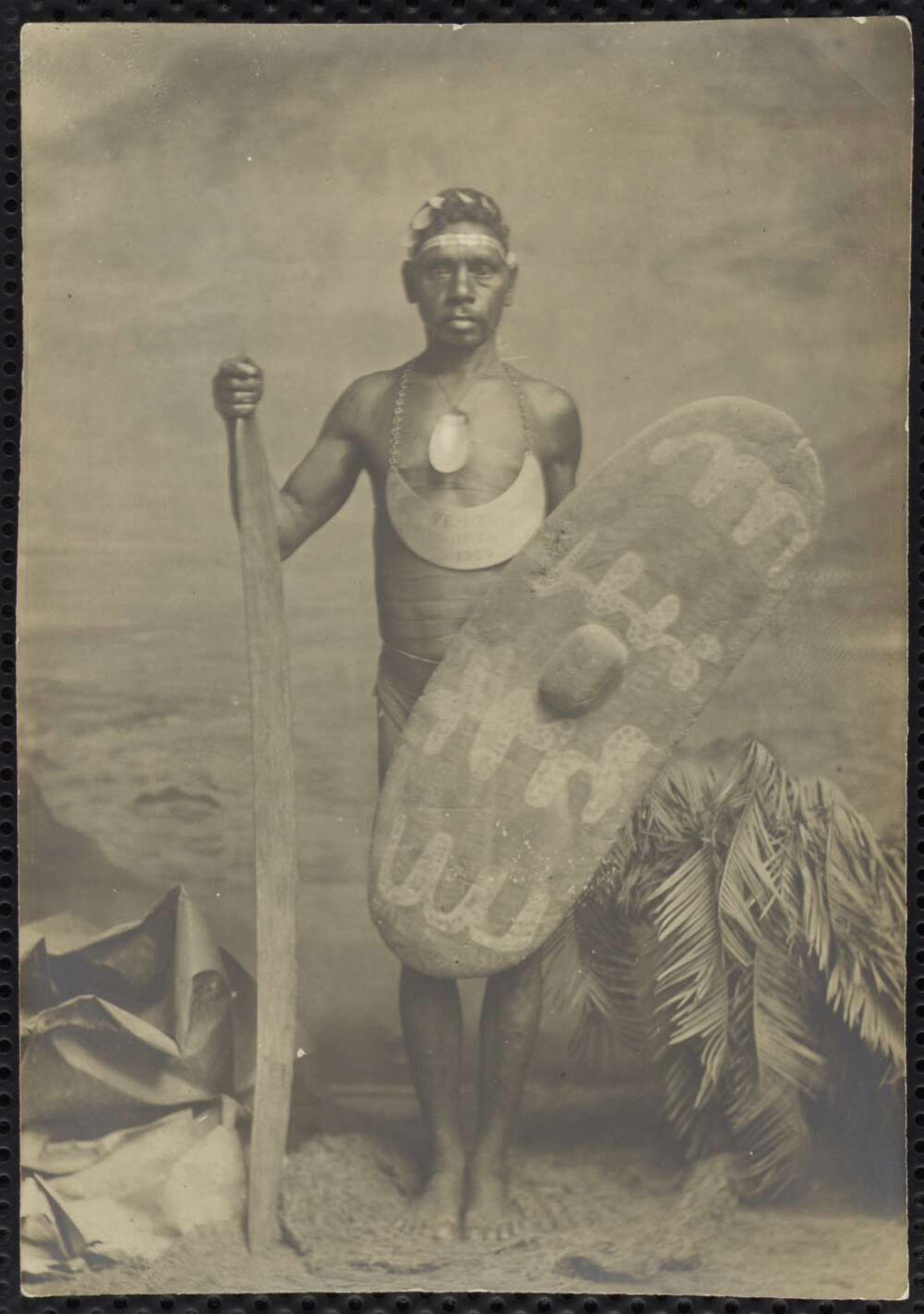 Black and white photograph of a man wearing a breastplate and carrying a shield and weapon. He is standing on a photo set that represents a beach with the sea in the background. - click to view larger image