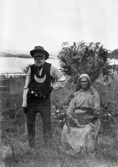 Black and white photo of a man standing and a woman sitting outdoors. The man is wearing a breastplate around his neck. - click to view larger image