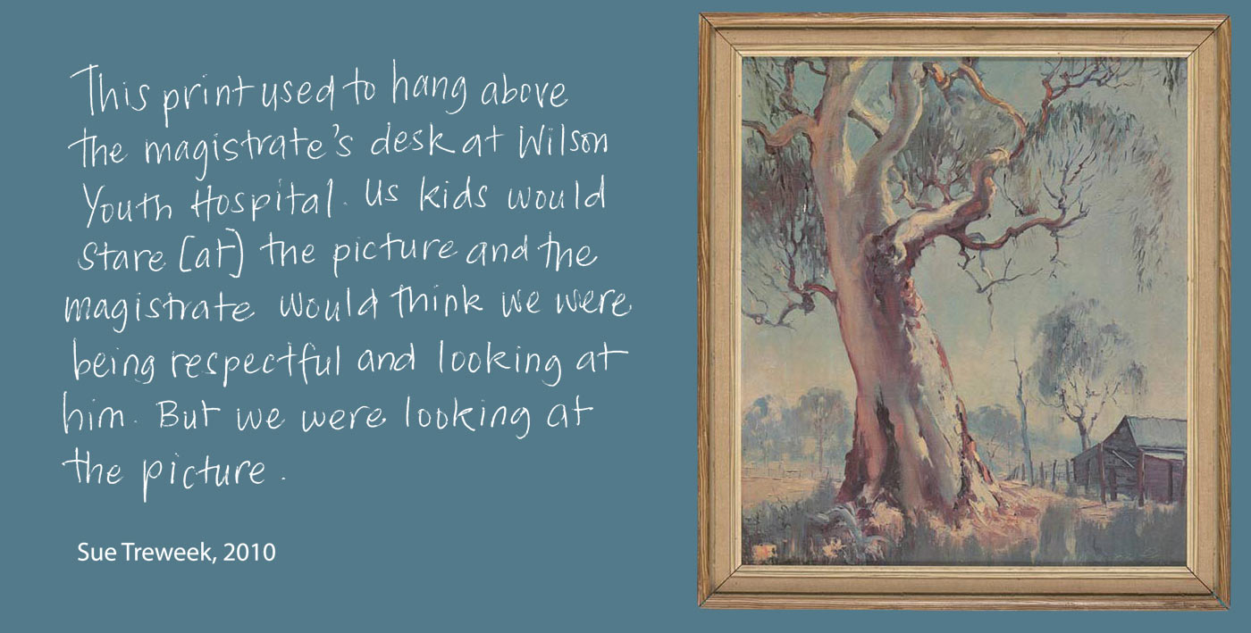 Exhibition graphic panel of white handwritten text on a grey background reads: 'This print used to hang above the magistrate’s desk at Wilson Youth Hospital. Us kids would stare [at] the picture and the magistrate would think we were being respectful and looking at him. But we were looking at the picture', attributed to 'Sue Treweek, 2010'. At right is a photo of a framed landscape painting showing a large central gum tree, a wooden fence at the right and part of a timber shed. - click to view larger image