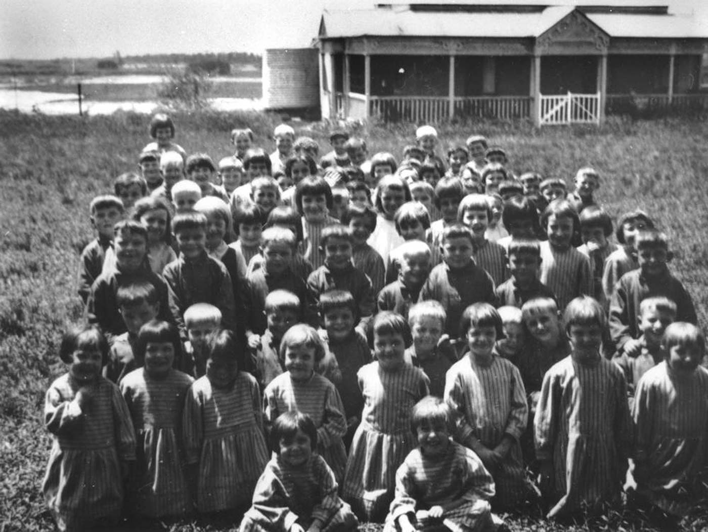 Black and white photo showing a group of about 50 children, sitting and kneeling on grass, with a wooden homestead in the background. The young girls wear striped dresses and the boys wear dark tops. Most of the children are smiling. - click to view larger image
