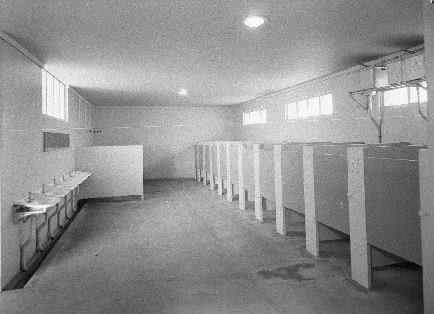 Black and white image showing a large white-walled room with a concrete floor. A row of six sinks lines the left wall and a row of partitions and doors lines the right. - click to view larger image