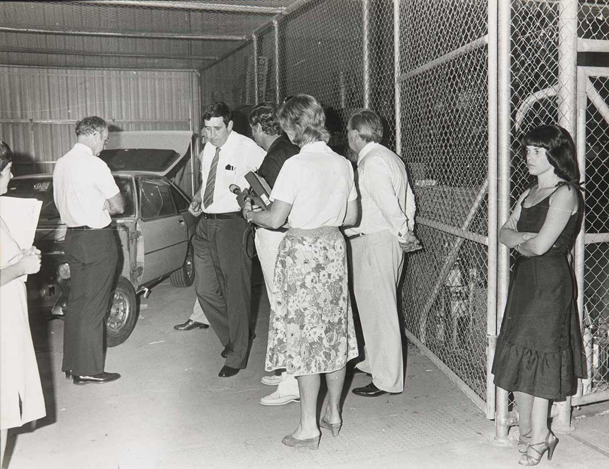 A woman is standing silently with arms crossed. A group of people who appear to be various officials and the media are congregated nearby.