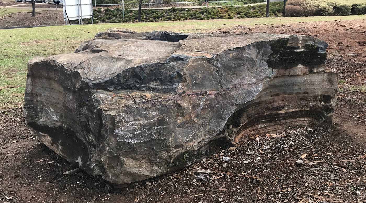 Large rock sitting in a park. The rock displays a rough surface with layers of sediment.