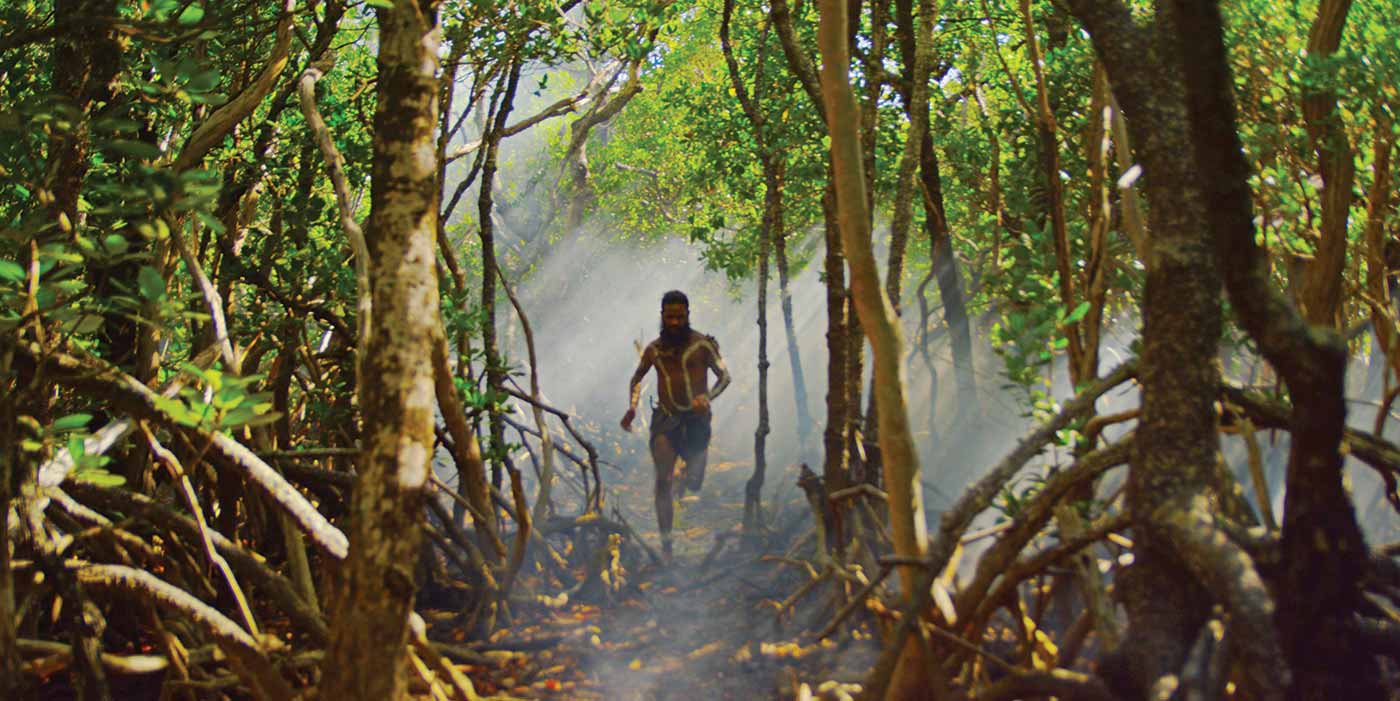 A man wearing animal skins and body paint runs through a forest.