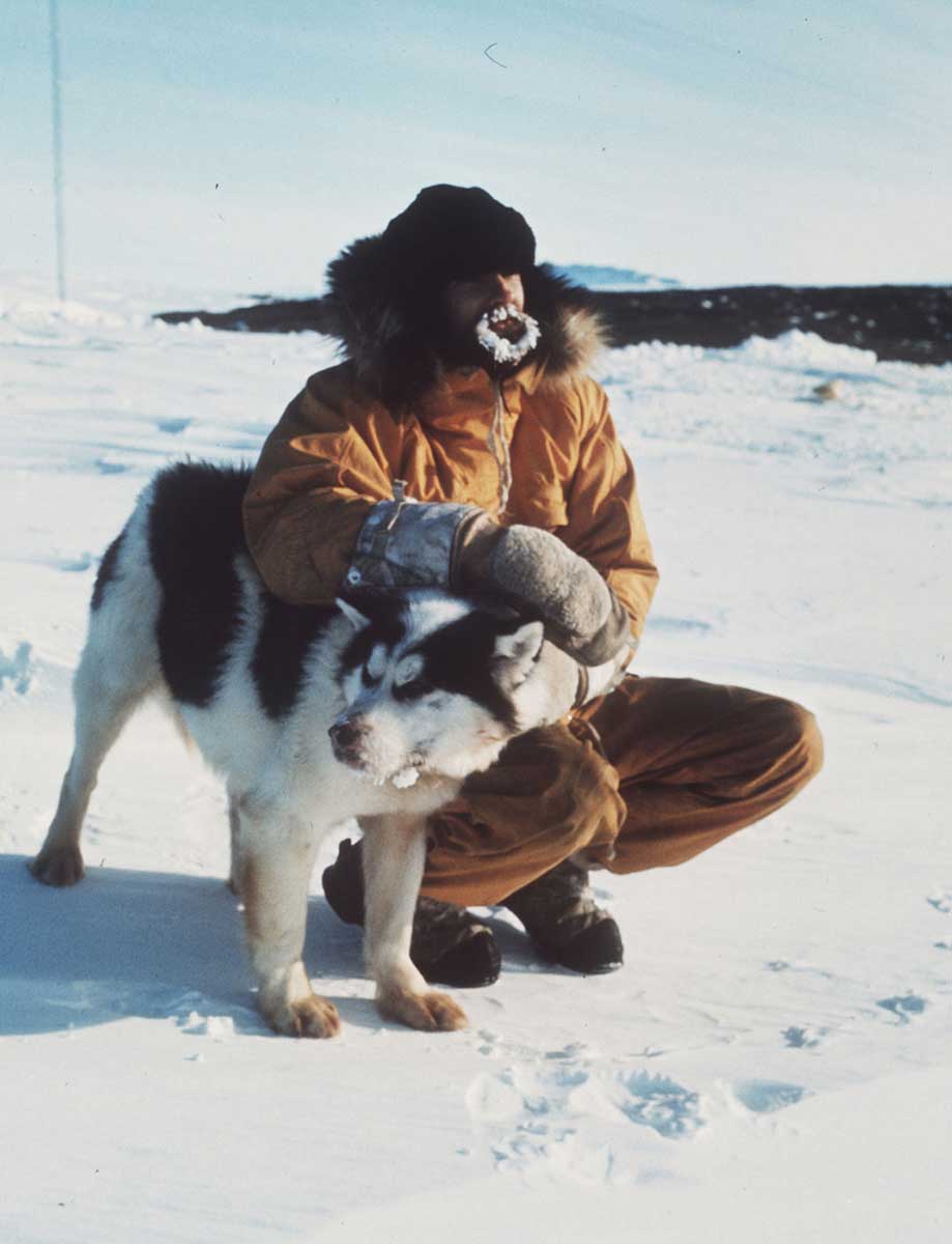 Colour photo of a man squatting with his arms draped over a husky. The man is wearing arctic gear and has a thick layer of ice that has formed over his facial hair. - click to view larger image