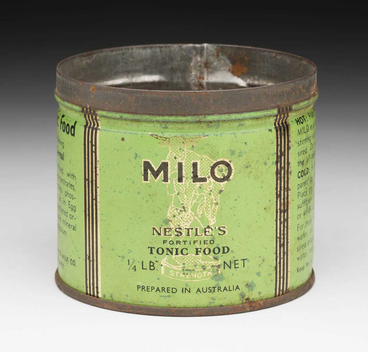 Round MILO tin without lid. The label is green, black and white and features an illustration of a bull being lifted by a man standing on a pedestal with the text ‘STRENGTH’ written on its side. on the label is the text ‘MILO / NESTLE’S / FORTIFIED / TONIC FOOD / 1/4 LB NET / PREPARED IN AUSTRALIA’. - click to view larger image