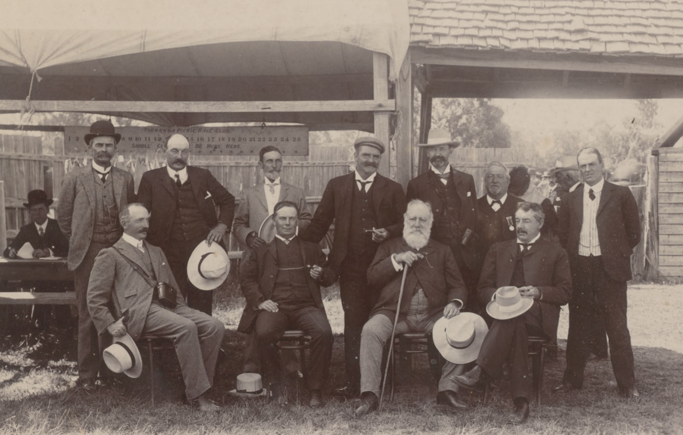 A black and white photograph that depicts four men seated in front row with Augustus Lucian Faithfull on the far right. Behind them seven men are standing with George Faithfull the first on the left. In the background a fence and open-sided building; on the left in background, one man seated at a table below a banner which reads 'Tirranna Picnic Race Club'. - click to view larger image