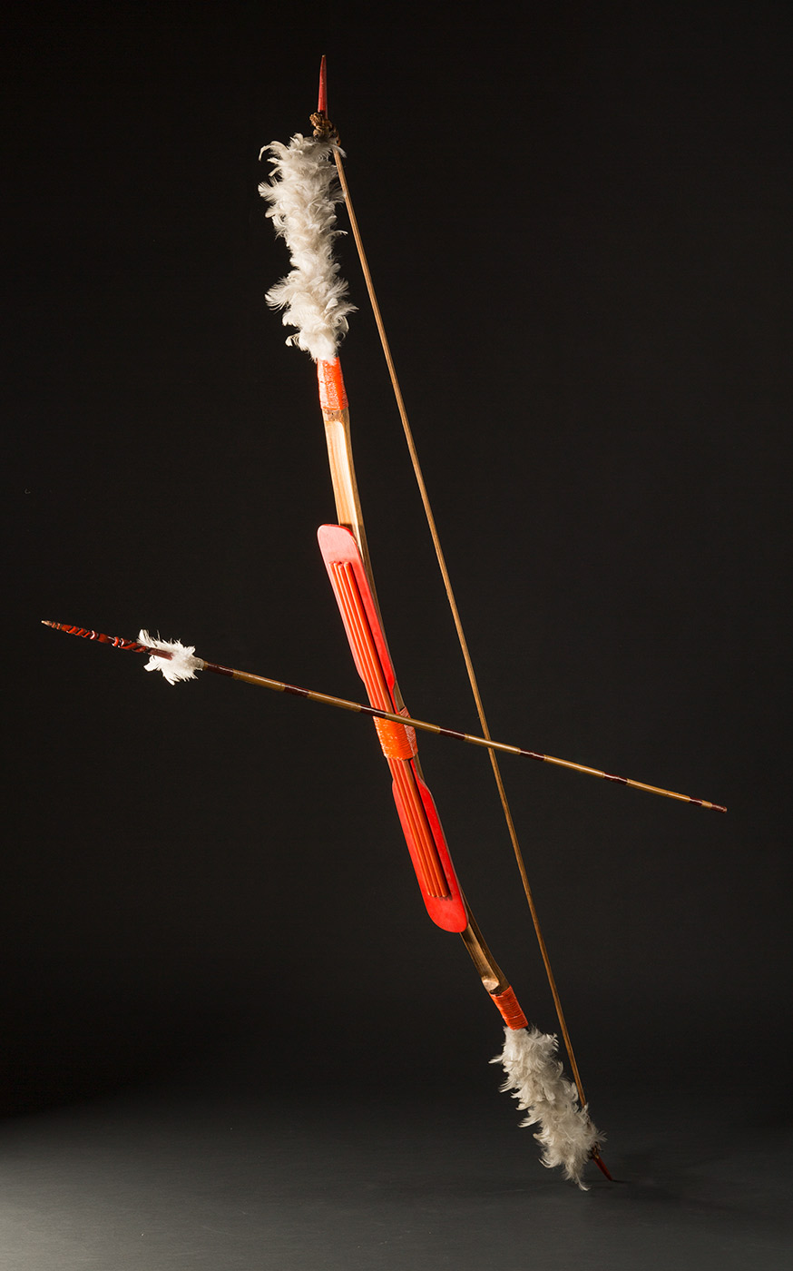 A bow and arrow set. The bow features a piece of wood with four cylindrical poles attached to the bowed wood and painted red. There are grey feathers attached to the ends bound with string also painted red. There is a thin piece of wood bound to the ends of the bowed wood with natural fibres. The arrow is attached to the bow with wire and features read bands the length of the shaft and red pigmented tip with carved twisted design and grey feathers attached. - click to view larger image