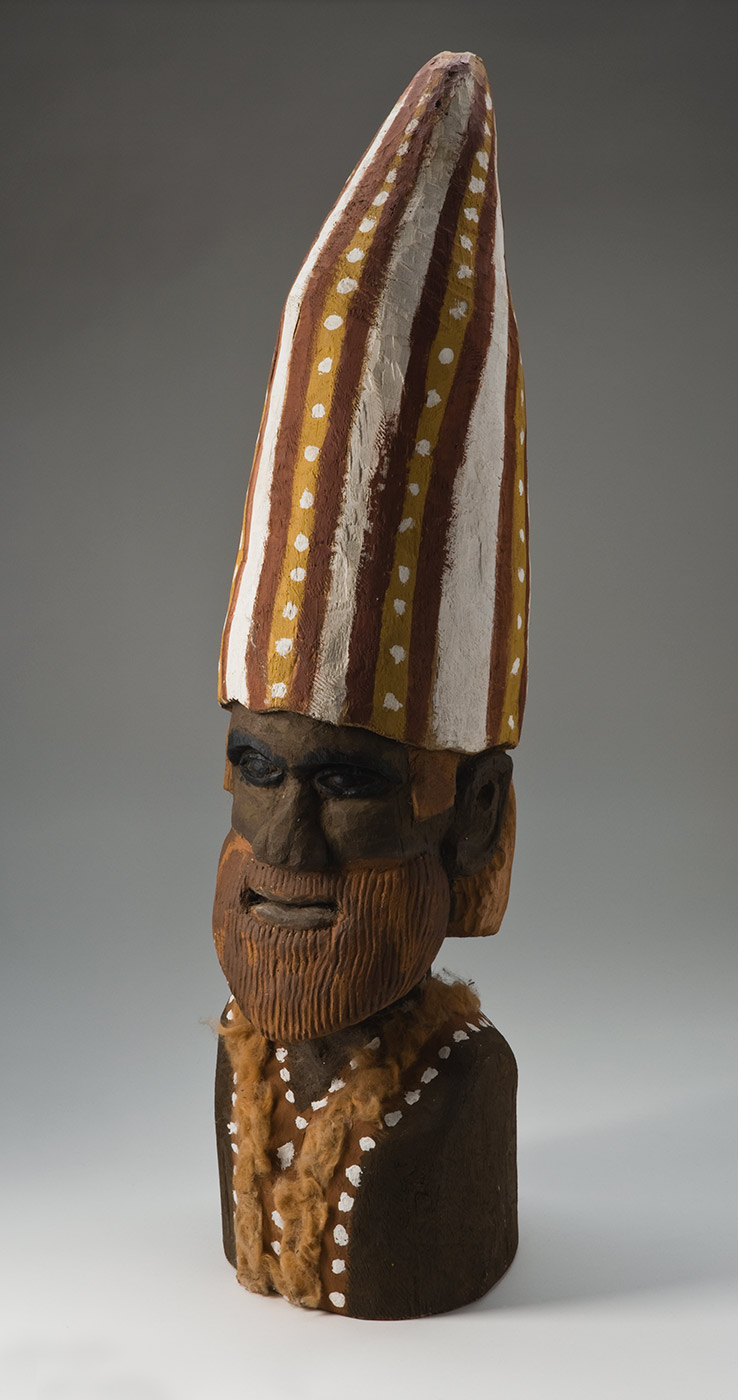 A carved and pigmented wooden bust of a human figure with a pointed hat and body decoration. The face and body of the figure are black-brown with the facial features deeply carved out of the wood, including an 