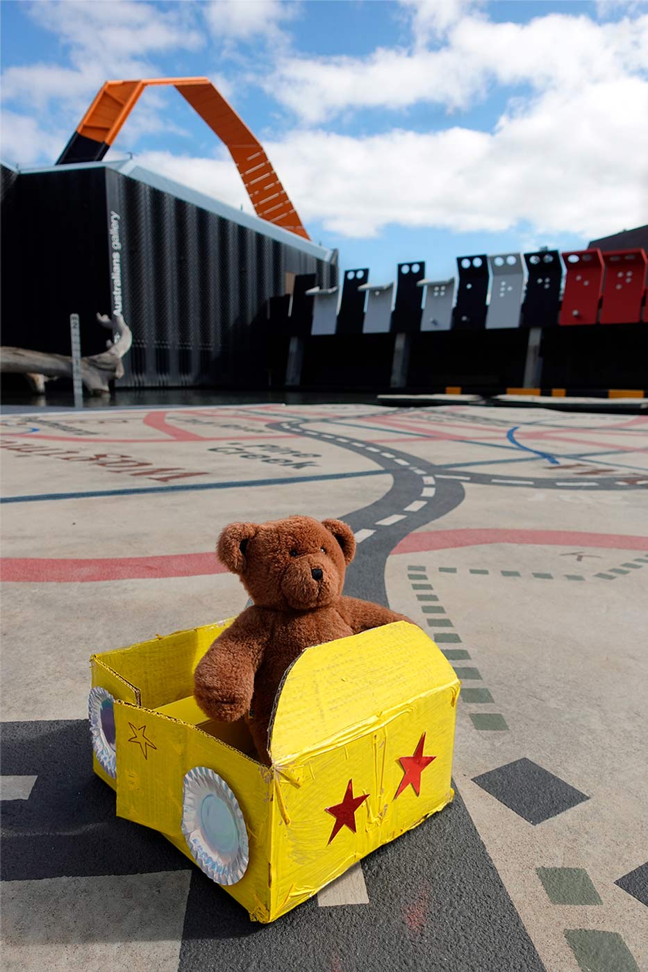 Colour photo of a stuffed toy teddy bear sitting in a handmade vehicle fabricated from cardboard and other found objects. The location is in the Garden of Australian Dreams at the National Museum of Australia.
