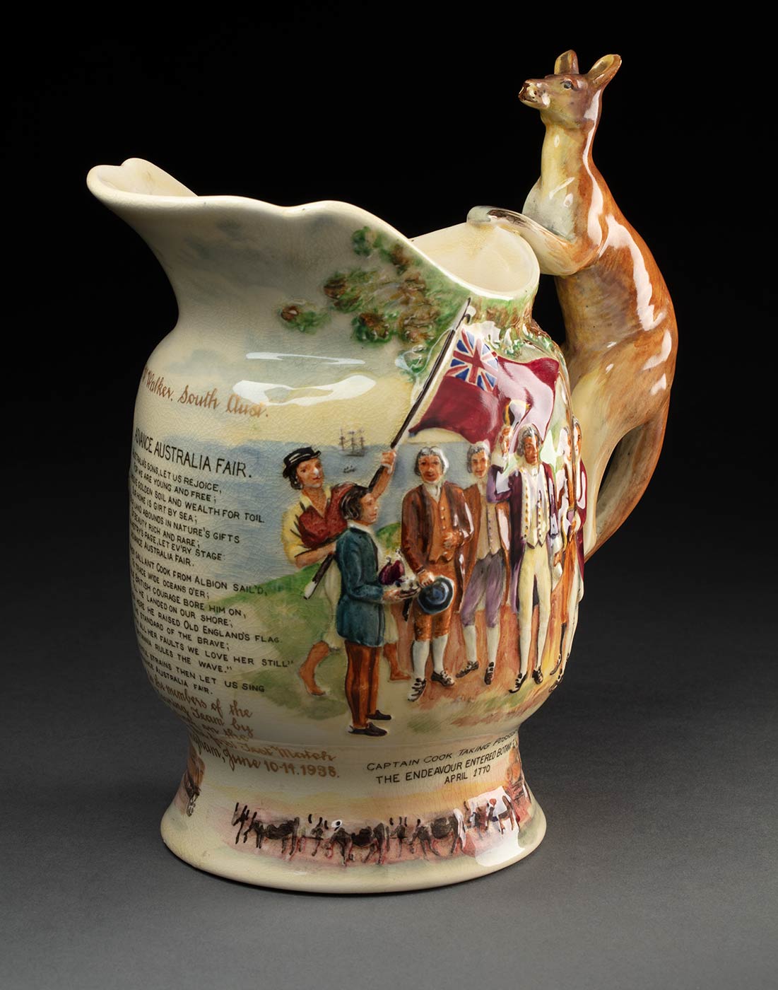 Fielding's Crown Devon earthenware 'Advance Australia Fair' musical jug which features relief moulding with a handle in the shape of a kangaroo. The jug has a painted scene of ships and a boat entering Botany Bay with Aboriginal men brandishing spears in their direction. Text below this reads, 'THE BLACKS THREW SPEARS AT COOK'S MEN.' There is also an image of Captain Cook raising his hat. He is surrounded by his men, one of whom is holding a red ensign. Text below this reads, CAPTAIN COOK TAKING POSSESSION. THE ENDEAVOUR ENTERED BOTANY BAY IN APRIL 1770'. The jug features the first and second verse of the poem 'Advance Australia Fair'. Text inscribed at the base in gilded lettering reads, 'Presented to the members of the Australian Touring Team by A. Ross. Fielding, on the occasion of the first test match at Nottingham, June 10.14.1938, C. W. Walker, South Aust.' Beside this is an untitled depiction of a drover's dray pulled along by a bullock team. The base is filled in with a wooden plate which has a winding mechanism set into it. The edge of the base has black text that reads 'CROWN DEVON / FIELDINGS / MADE IN ENGLAND'. - click to view larger image