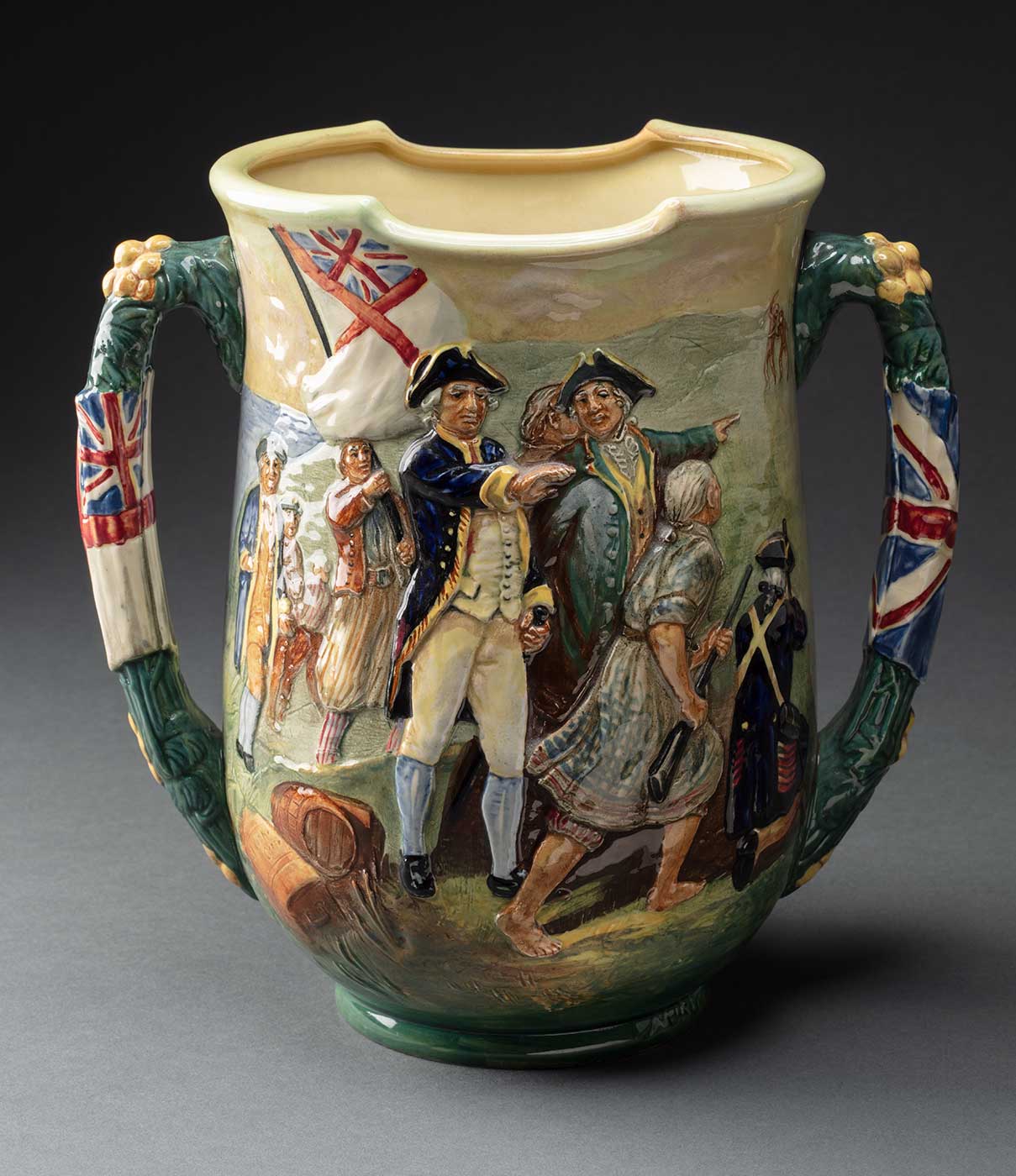 Royal Doulton loving cup (with two handles) depicting a scene of the early Australian colony. One one side Captain Cook and eight other men are shown in relief, with two figures of Indigenous men painted in the background. On the other side a sailing ship and two rowboats are shown with four figures on the shore in the foreground and several figures with the rowboats. Flags are depicted on the handles. The underside is stamped with 'Captain Cook / The Greatest Navigator / ... / Through storm & shine / alone on unknown - / seas. The Endeavour / anchored in Botany Bay / ub tge tear if iyr Kird / 1770' and 'The production of this cup / is limited to 350 / no. 215'. The trademark stamp reads 'Made in England / Royal Doulton / Registered / in Australia / No. 10610'. - click to view larger image