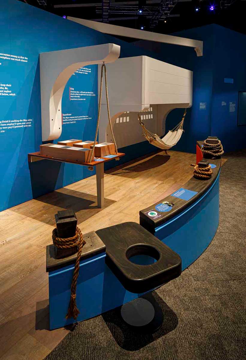 Interior view of a museum exhibition with a canvas hammock, ropes tied around bollards and a timber toilet seat. - click to view larger image