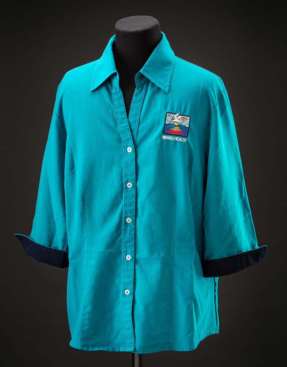 A turquoise shirt that features navy blue lining, white buttons and an embroidered patch featuring an ocean scene with a seagull, two Dolphins and the sun on the horizon. Underneath is embroidered text: ‘MIWATJ HEALTH’. - click to view larger image