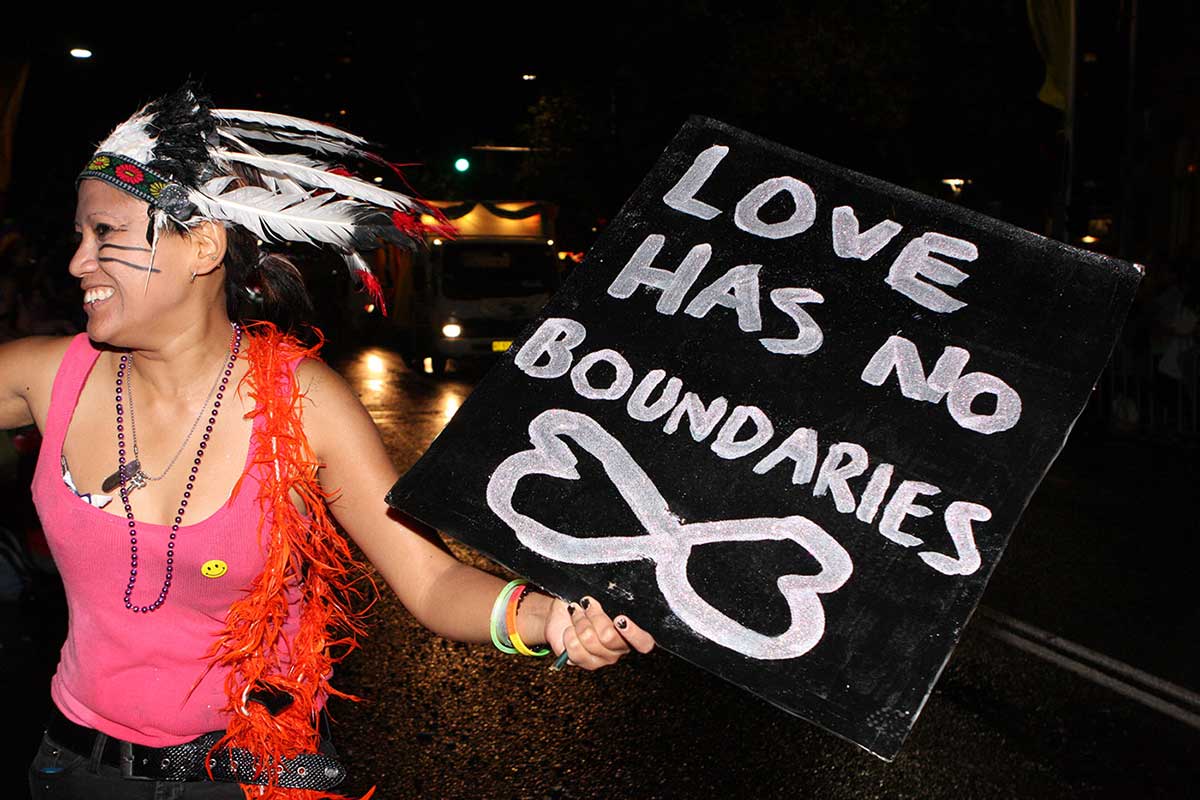 Woman in feathered headdress holding placard saying 'Love has no boundaries'.