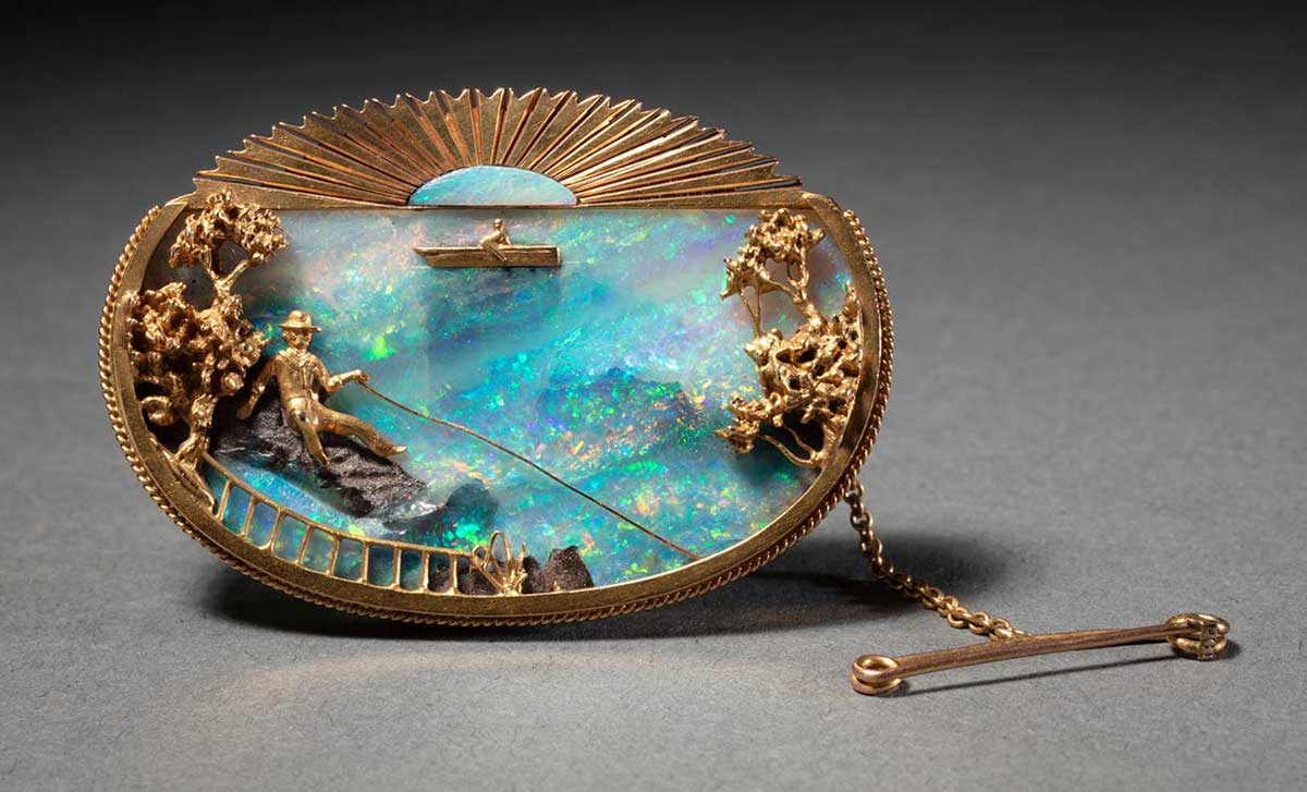 A gold oval brooch set with a blue opal and depicting a man sitting on a rock fishing in the foreground with trees on either side and a fence. There is a boat in the background and a sun motif at the top. - click to view larger image
