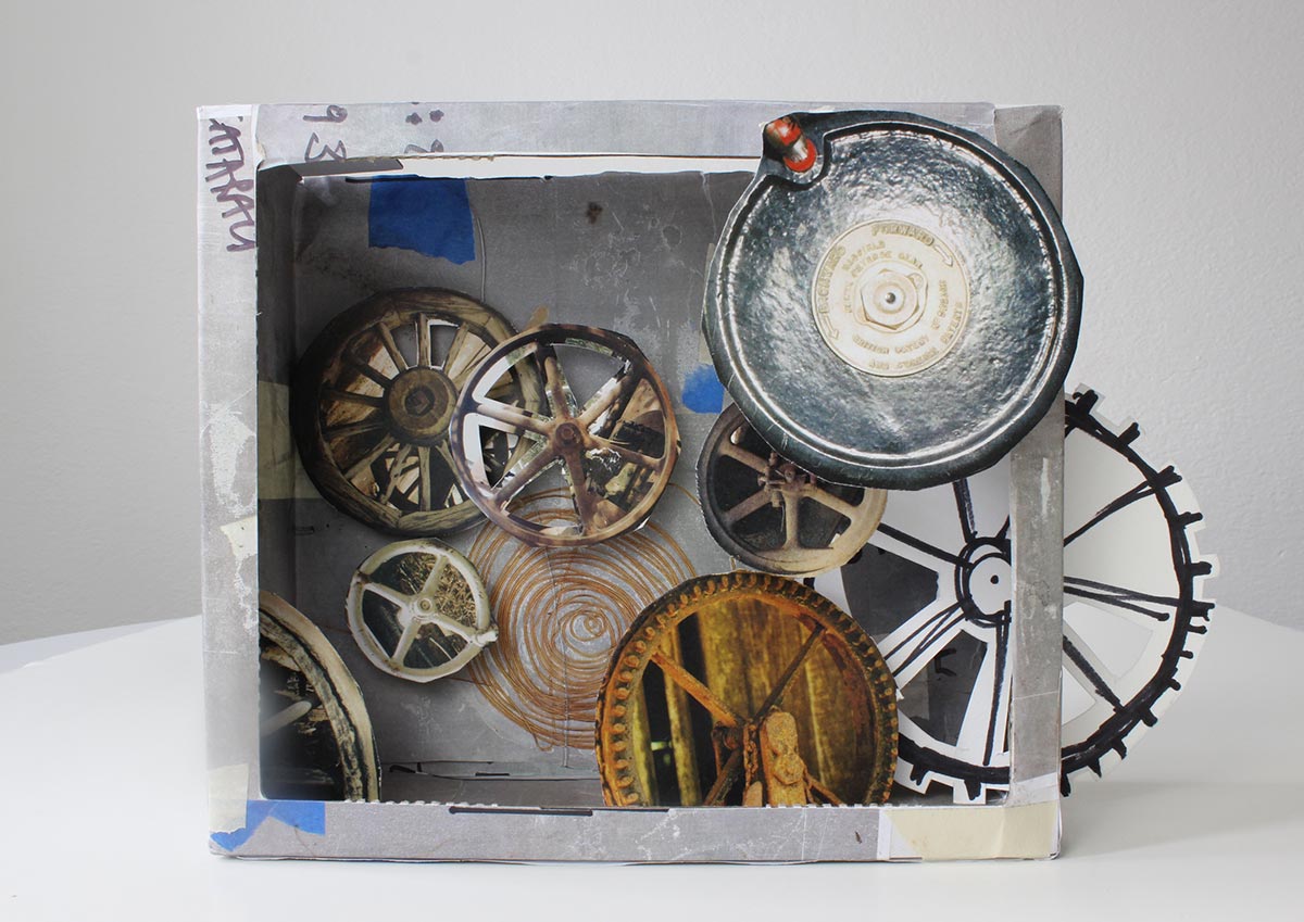 Diorama constructed from cardboard and various other materials. The interior of the box features cut-out colour images of round mechanical objects.