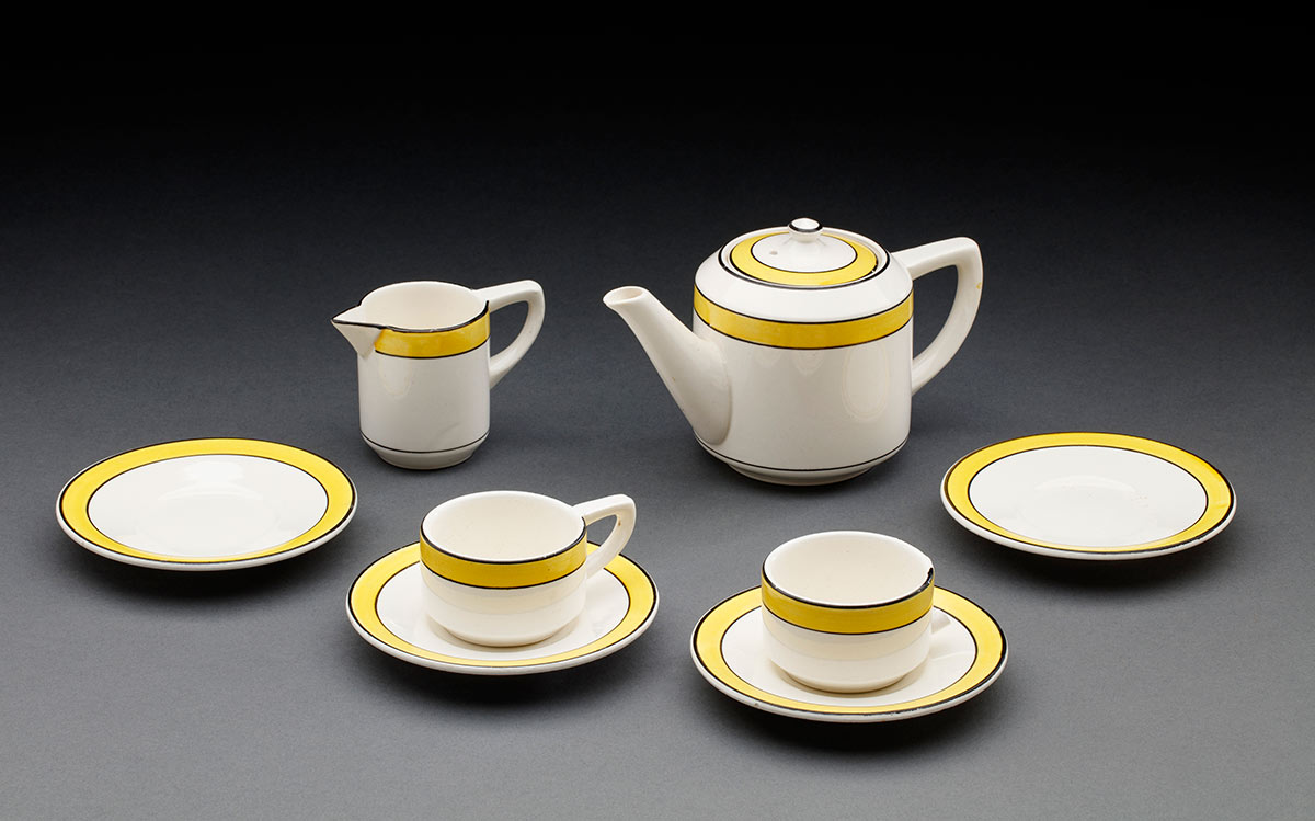 A white ceramic transfer printed toy tea set. It includes a tea pot, a milk jug, two tea cups (one is missing the handle) and four saucers. There is a yellow stripe around the outer edge of each piece and there are black lines outlining the yellow stripes. - click to view larger image