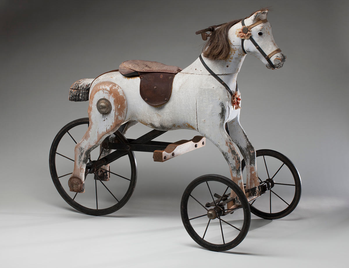 A white wooden horse tricycle mounted on a metal frame, with a metal and wooden rear wheel. The wooden horse has painted on black eyes, moulded nose, mouth and ears, a horse hair mane, and leather bridle and saddle. The steering handles are linked to two small steerable wheels through the neck and the forelegs, and rotating hind legs are attached to the larger rear wheel. The paint is worn, chipped and cracked. - click to view larger image