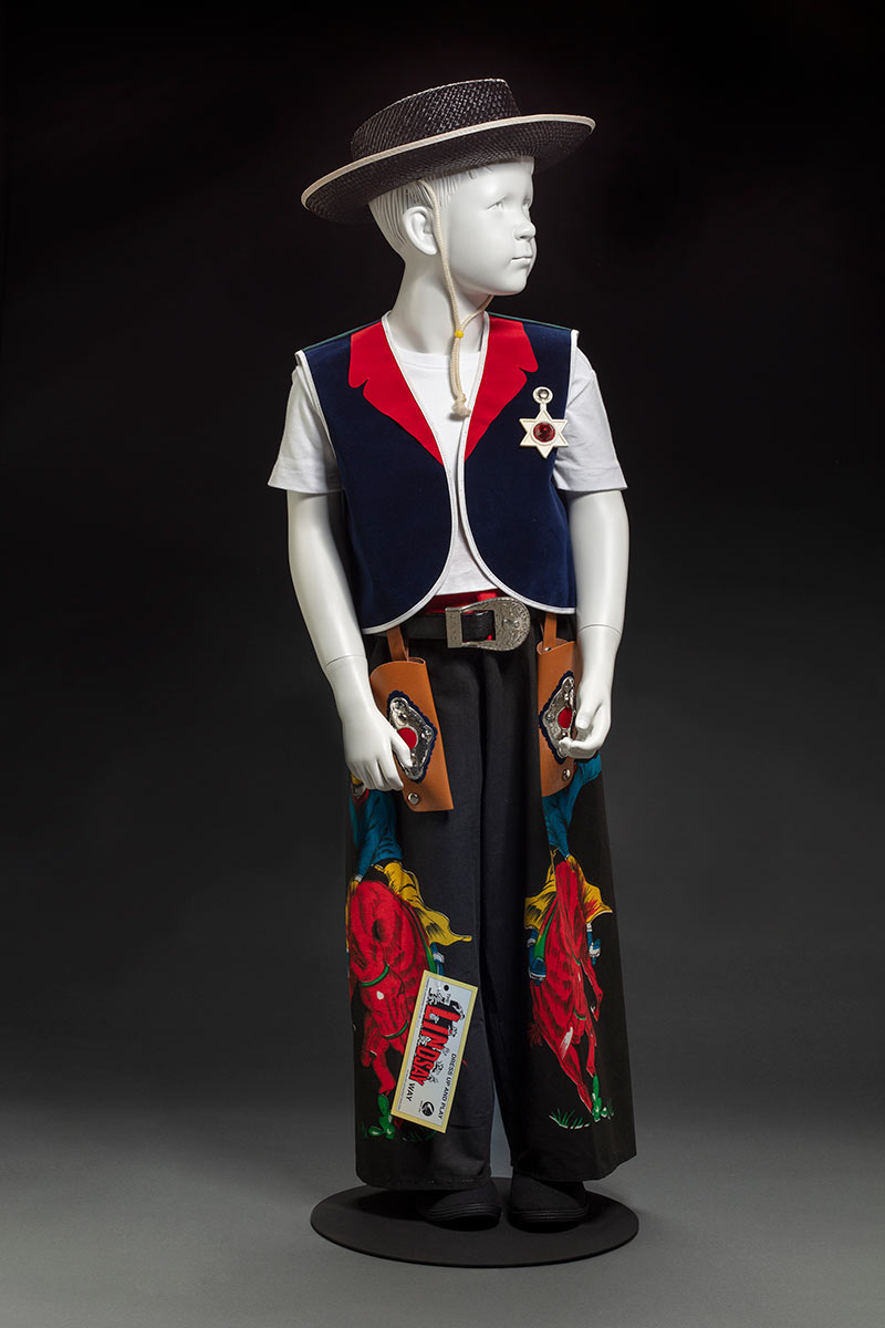 A child's cowboy costume comprised of a red and blue vest with a green and blue tartan back, and a pair of black and red pants featuring images of a person riding a bucking horse. Attached to the pants is a label attached. - click to view larger image