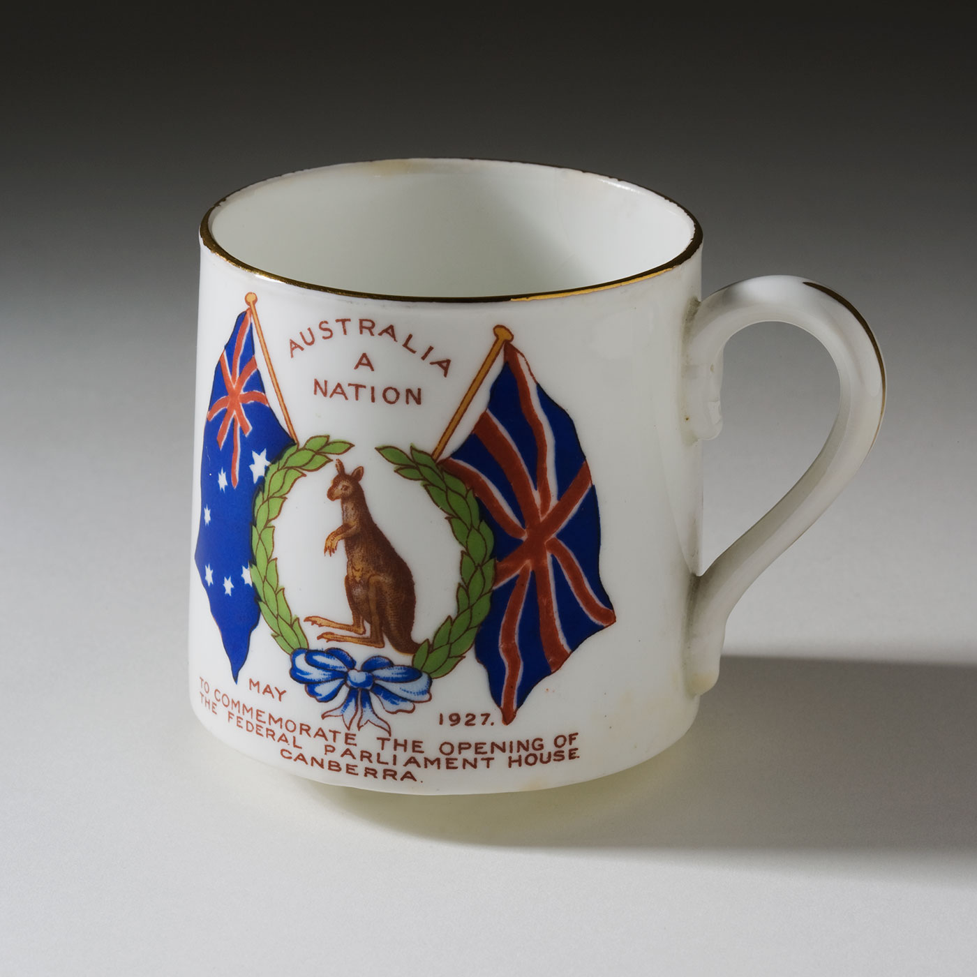 White mug with gold rim showing a kangaroo in a garland of leaves. 'Australia A Nation' is printed at the top, between the Union Jack and Australian flags which hang at either side of the illustration. 'May 1927, To commemorate the opening of the federal parliament house, Canberra' is printed at the bottom of the cup. - click to view larger image
