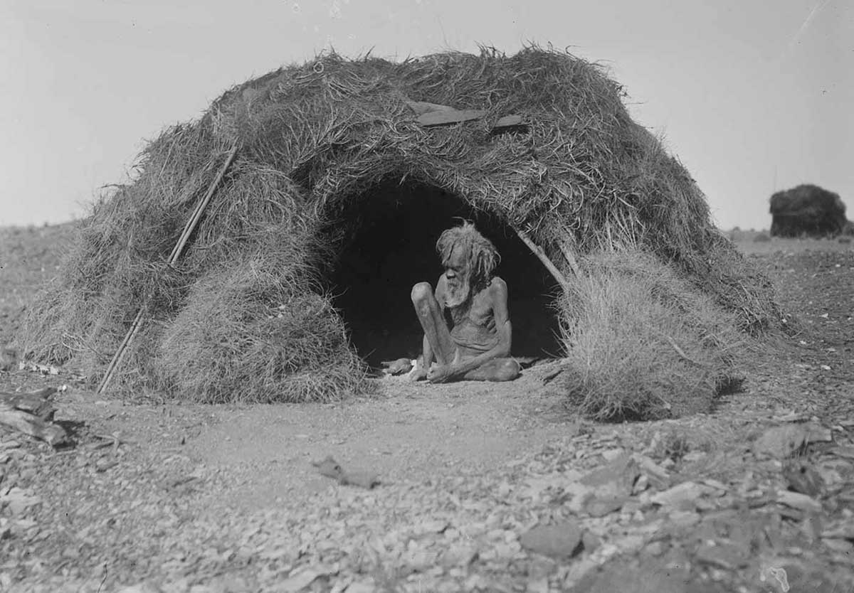 Elderly Aboriginal man sitting in front of a hut, Northern Territory, August 1920. The hut is decked with porcupine grass and stands about the same height as an adult. It has a domed form with a simple opening providing access. The man sits on the ground in the opening. He has his left leg folded under him and his right leg bent so that his right knee is at chin height. He has long hair and a full beard. His body is very thin and gaunt. the ground directly in front of the opening is bare. In the foreground are rocks and stones. A large bundle of porcupine grass sits on the ground to the right of the hut opening. In the right background can be seen another hut. The horizon can be seen in the left background. The landscape around the huts is flat and featureless.
