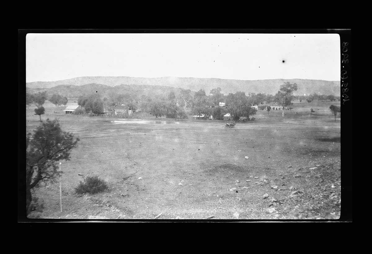 Alice Springs, Northern Territory 1920. The photo has been taken from a low hill near the town. Trees in the middle distance obscure some buildings. Individual buildings can be seen in the left and right middle distance. Beyond the trees and houses is a low range of hills. In the far background is a larger range that runs across the entire image. Cloudy white spots on the image suggest some sort of damage or ageing in the photographic emulsion. - click to view larger image