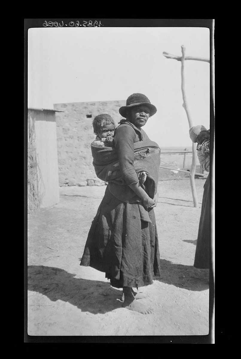 An Aboriginal woman with an infant being carried on her back, Denial Bay, South Australia 1920. The woman wears a dress and hat; she is barefoot. She stands facing the right side of the image with her head turned toward the camera. The infant sits on her back in a carrier made from material wrapped around the woman's body. Another infant being carried on a woman's back can just be seen on the right side of the image. - click to view larger image