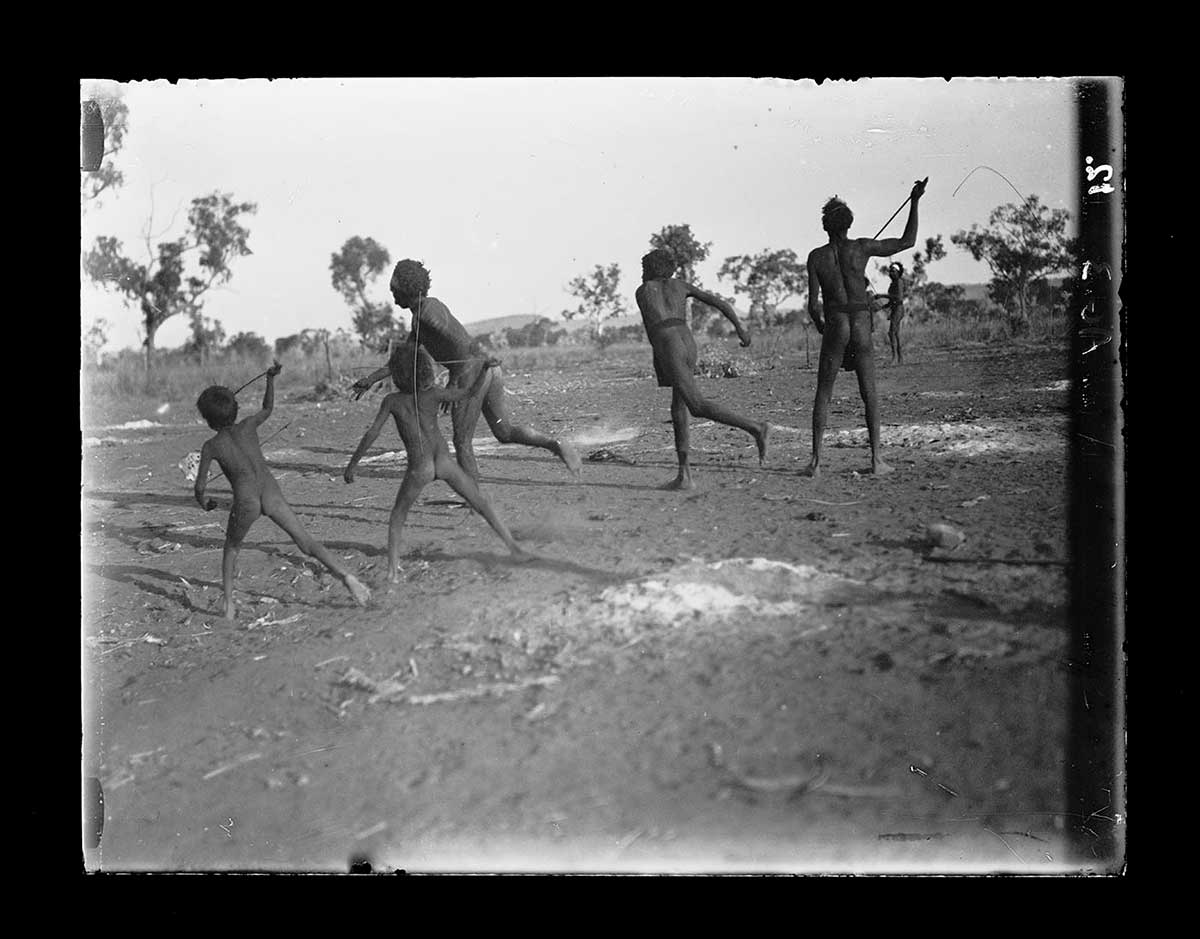 Aboriginal men and boys playing a customary game, Humbert River station, Northern Territory 1922. Three men and two boys are participating. They all have their backs to the camera; the boys are closest to the camera on the left of the image. A rolling bark disc is out of shot to the left. The two boys and two of the men are throwing spears at it. The third man stands with a spear held at head height in his right hand. Another man watches the game from the right background. - click to view larger image