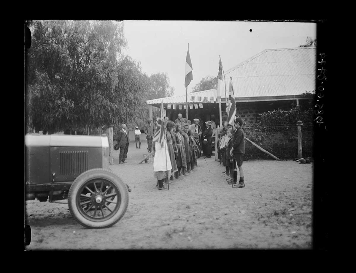Guard-of-honour for the Governor of South Australia, Alice Springs, Northern Territory 1923. Two rows of Aboriginal and non-Aboriginal children face each other about two metres apart. The children are standing outside a single-storey building with a verandah and corrugated iron roof. A child toward the camera end in each row holds a Union Jack flag on a pole. Toward the rear of each row, close to the building, what may be the Northern Territory and South Australian flags are on taller poles. Several adults are in the background waiting for the Governor. The front half of a car is seen in the left foreground. - click to view larger image