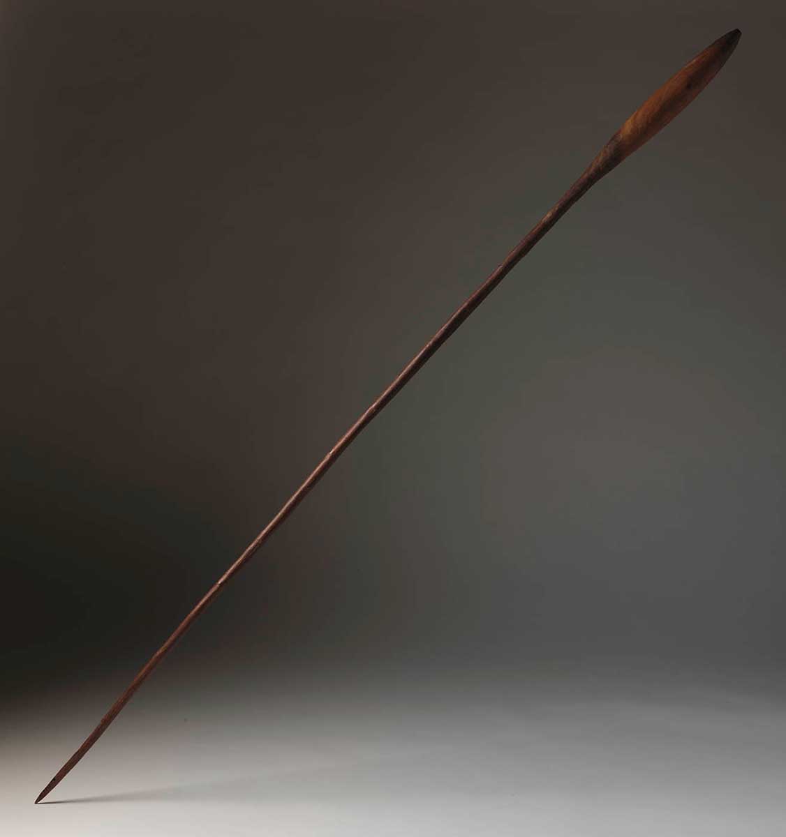 A long, cylindrical smoothed wooden stick. It has a sharp point at one end and a slightly bulbous section at the other end, about one fifth of the stick in length. The bulbous section is slightly lighter in colour to the rest of the stick. The stick's surface is smooth and shiny, as though it has been polished or covered with a natural lacquer. - click to view larger image