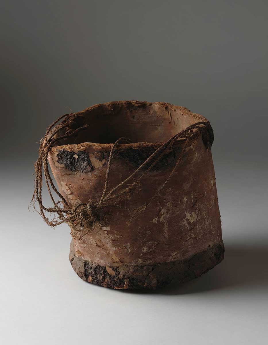 A bark container with a rough bucket form. The base has been attached to the sides with split cane and natural resin. String made from natural fibres is attached at opposite sides of the container's top. They form a loose handle which hangs over the container's side. Small areas of light pigment are on the side facing the camera. - click to view larger image