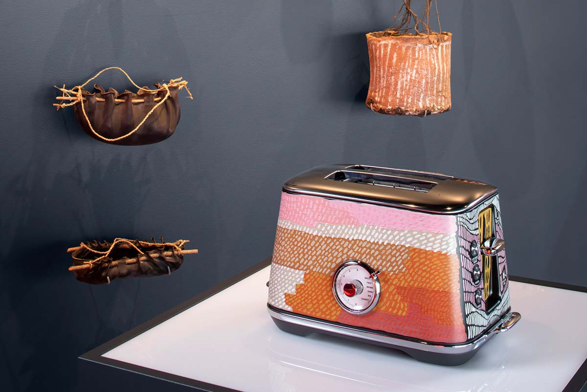 A toaster with a painted design on display in a gallery space. On the wall behind are various hand-crafted vessels on display. - click to view larger image