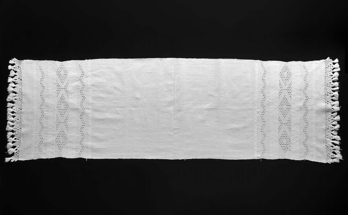 Long rectangular piece of off-white woven fabric, with crocheted edging and tassels at either end. The outer thirds of the long fabric are decorated with perforated zigzag and diamond patterns. - click to view larger image
