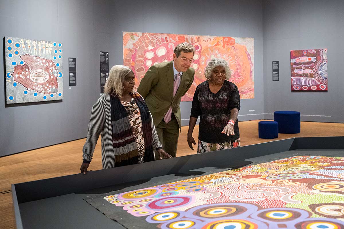 A man a two women inspect a large canvas painting in an exhibition space.