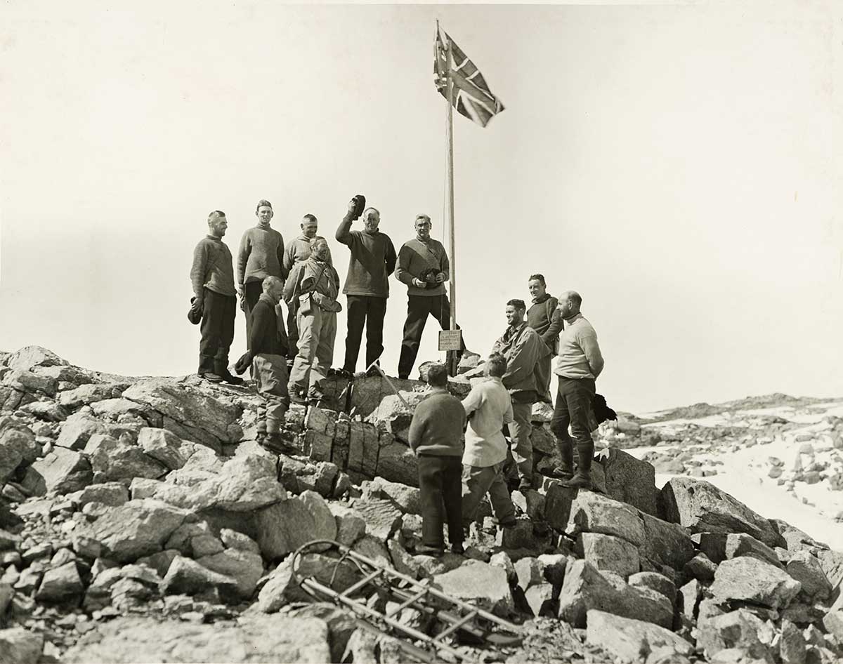 A group of men stand on a rocky mound, beside a flag pole flying the Union Jack. - click to view larger image