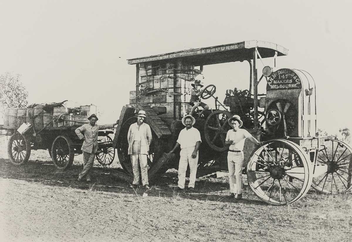 Black and white photograph of four men standing in front of a large stationary tractor with a young child sitting at the steering wheel. - click to view larger image