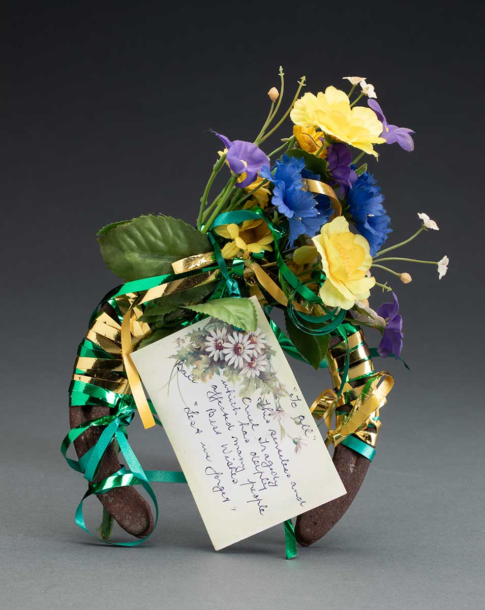Rusty metal horseshoe covered in a bouquet of cloth and plastic flowers. The yellow and purple flowers are tied to the horseshoe with strands of gold and green ribbon. A card attached reads '