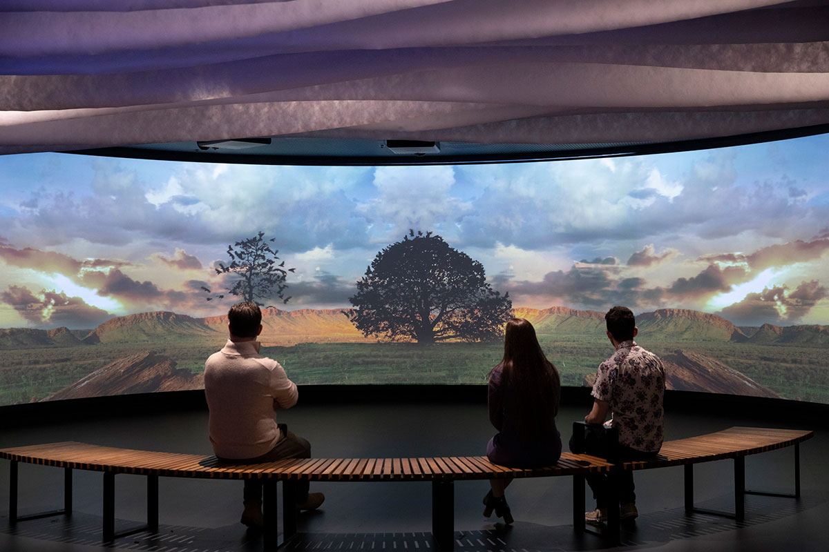 Three visitors sit watching a large digital screen with a projection of a vast landscape.